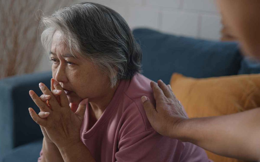 Gray-haired Asian old woman sitting on sofa feeling depressed while talking to psychologist. He placed a hand on her shoulder to cheer, concept mental health in old age